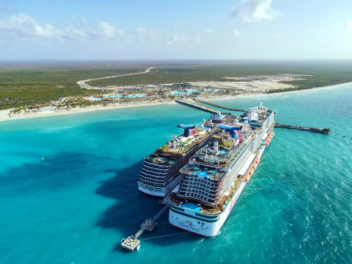 Two large cruise ships, including the Mardi Gras, are docked side by side at Celebration Key’s pier on clear turquoise water. A coastal resort, sandy beaches, and lush greenery grace the background under a mostly clear sky.