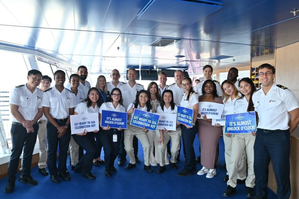 A group of cruise ship crew members in uniform and casual attire stand together indoors, some holding blue and white signs with messages like “It’s Almost Unlock O’Clock,” gearing up for a Grand Bahama locals tour at SEA Celebration Key.