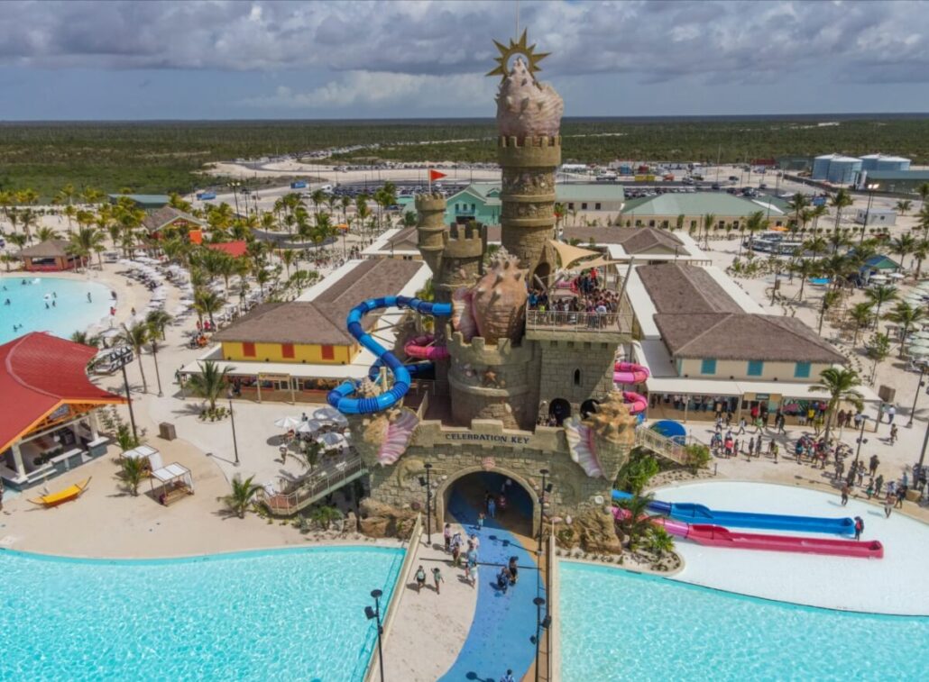Aerial view of a large water park on Grand Bahama featuring a castle-themed structure with slides, surrounded by pools, palm trees, and various buildings; people are visible enjoying the attractions under a partly cloudy sky.