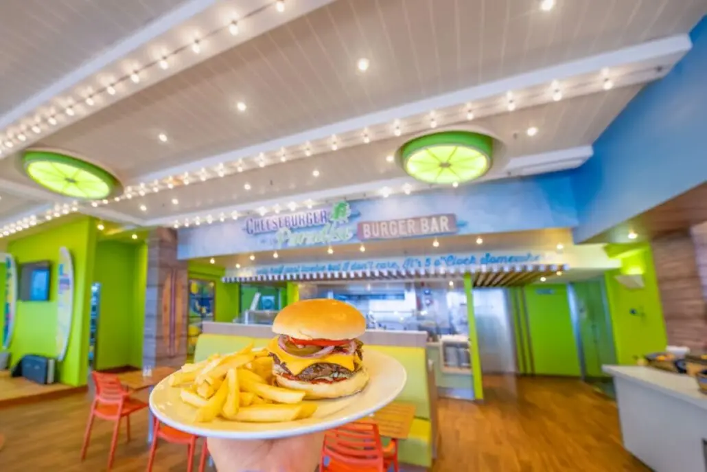 A hand holds a plate with a cheeseburger and fries in a brightly lit restaurant with green accents, red chairs, and surfboard decorations. The counter and a sign reading "Burger Bar" add to the fun Margaritaville at Sea vibe in the background.