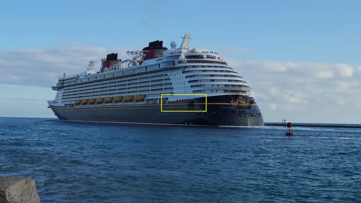 A large Disney Cruise ship with multiple decks and red funnels sails near a harbor under a partly cloudy sky. The white vessel with dark lower decks glides through calm water, offering excitement for every 5-year-old on board.