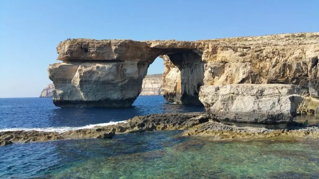A natural limestone arch known as the Azure Window rises above the blue sea on Gozo, with rocky formations and shallow water in the foreground, under a clear sky—a must-see for any tourist seeking hidden gems.