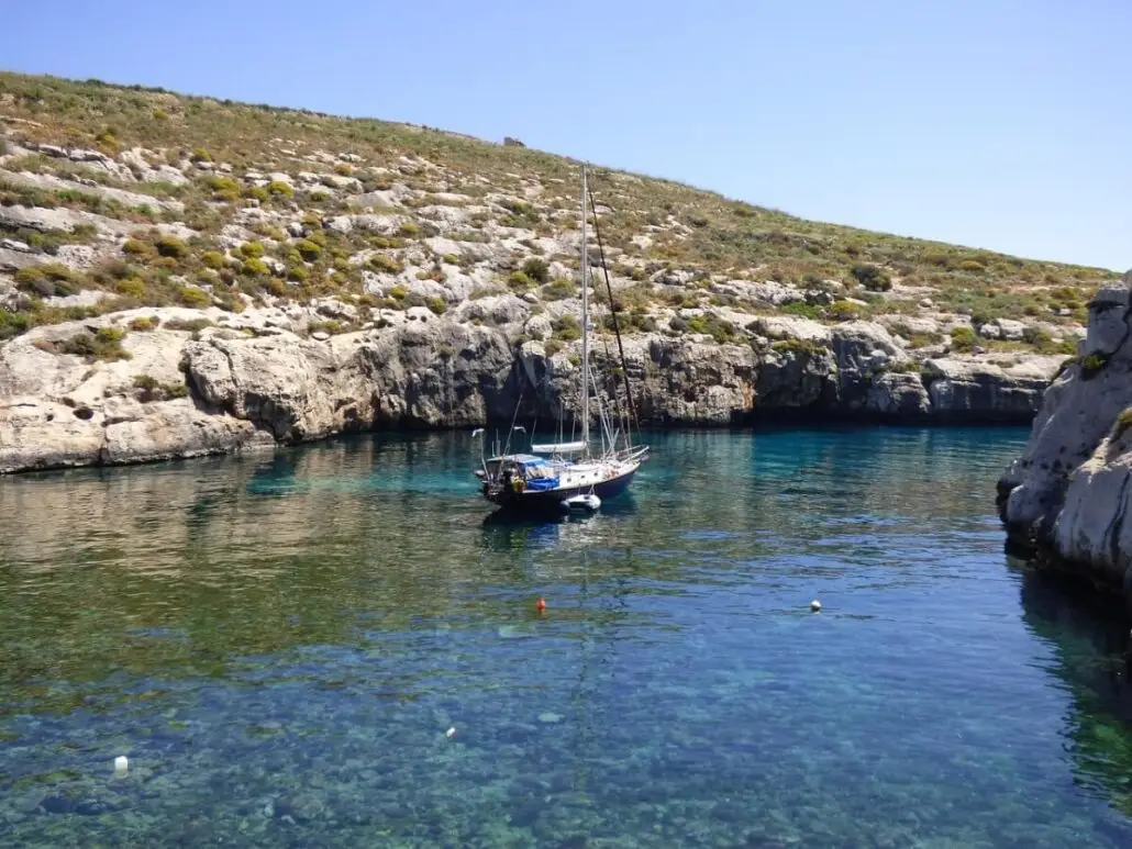 A small sailboat is anchored in a calm, clear blue cove—one of the hidden gems Gozo has to offer—surrounded by rocky cliffs and sparse vegetation. The water reveals underwater rocks along a rugged, uneven shoreline under a clear sky.