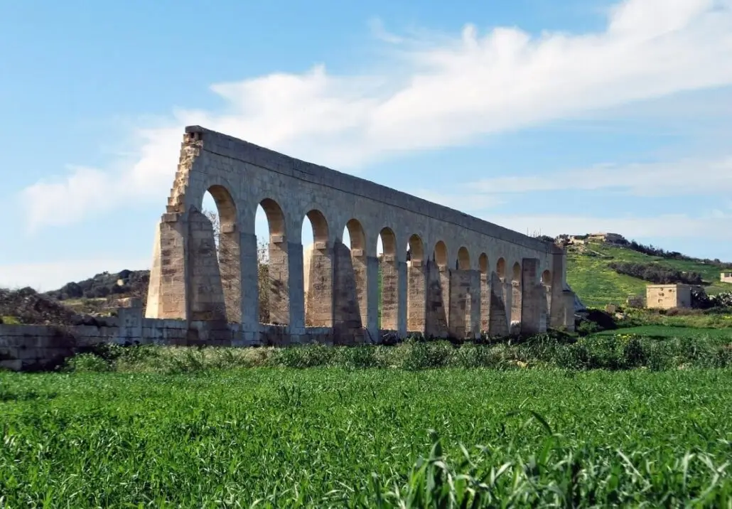 A stone aqueduct with multiple arches stands in a grassy field under a blue sky with wispy clouds—one of the hidden gems in Gozo. The aqueduct stretches into the distance, framed by green hills and a few small buildings in the background.