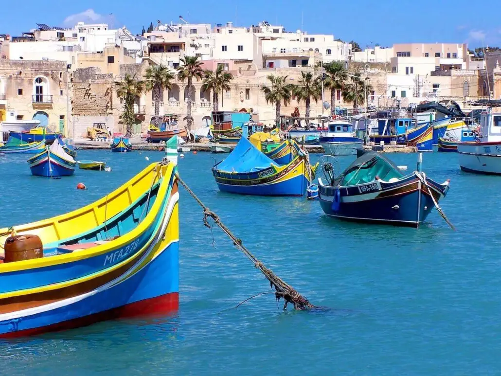 Colorful fishing boats float on blue water in a Gozo harbor, with a row of palm trees and white buildings under a clear sky—one of the island’s tourist hidden gems waiting for you to explore.