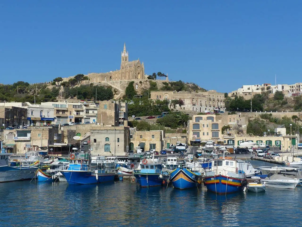 A harbor with several colorful boats docked in calm water, with stone buildings and a large church on a hill in the background under a clear blue sky—one of the hidden gems Gozo has to offer for those who love to explore Gozo’s authentic charm.