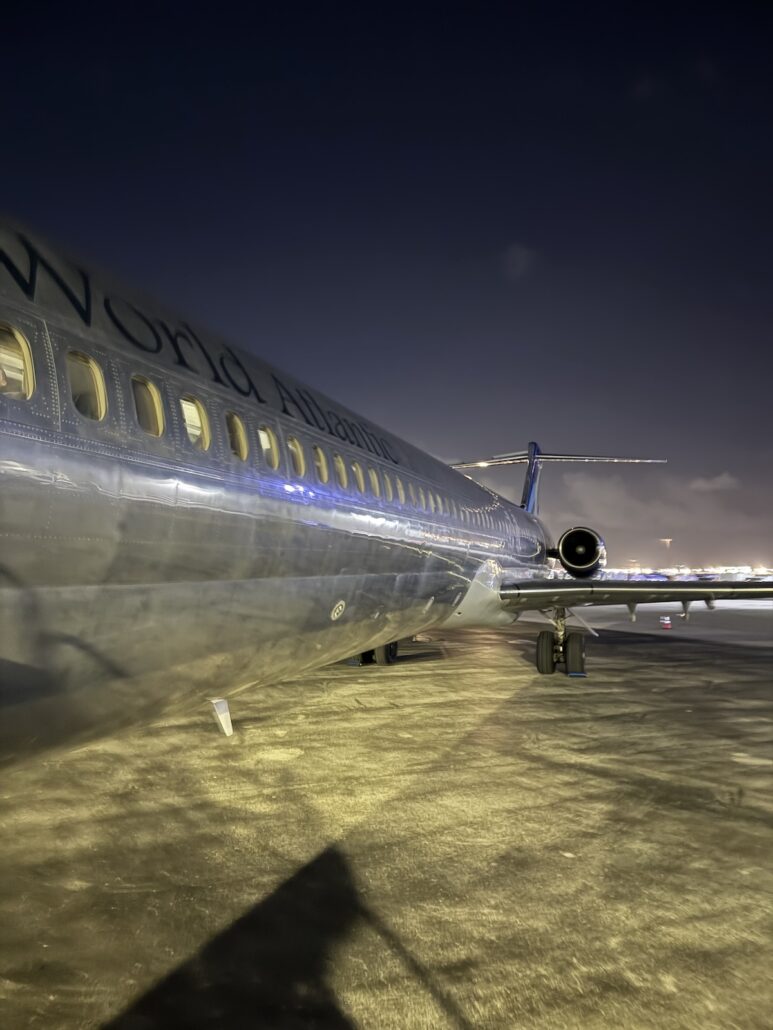 A commercial jet is parked on the airport tarmac at night, its metallic fuselage gleaming under runway lights—a behind-the-scenes glimpse of preparations for a grand opening celebration.