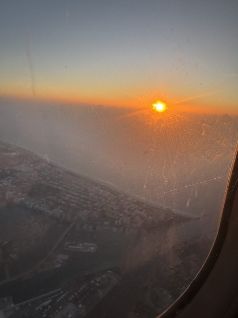 Photo taken from an airplane window shows a sunset above a coastline, with buildings and water visible below; the window has visible smudges and scratches, capturing a unique behind the scenes view from above.