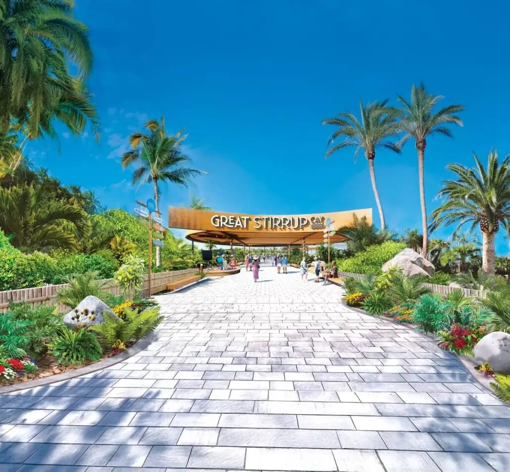 A wide stone pathway lined with tropical plants leads to an entrance sign that reads “Great Stirrup Cay,” Norwegian Cruise Line’s private island paradise, under a clear blue sky with palm trees and a few people strolling along the path.