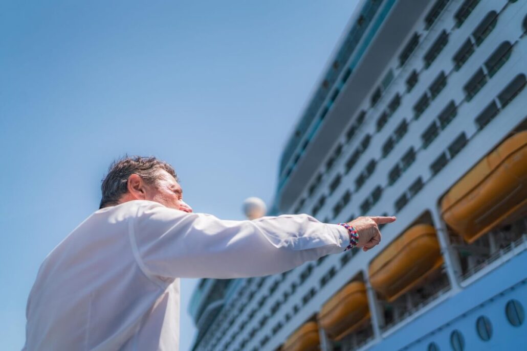 A man in a white shirt is standing outdoors, pointing towards a large cruise ship. The ship's side, with multiple windows and lifeboats, is visible against a clear blue sky, hinting at a possible confrontation involving the Mayor of Nice.