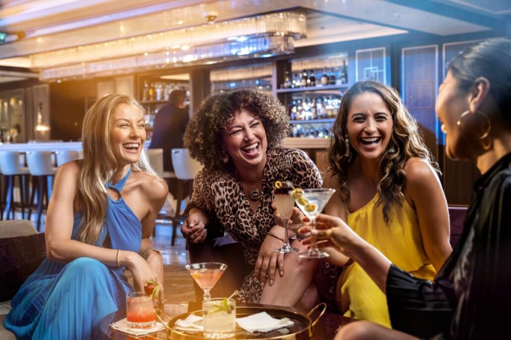 Four women sit together in a bar or lounge on Princess Cruises, smiling and laughing while holding cocktails. There is a tray with drinks on the table, hinting at the indulgence of the Premier Package, with a stylish bar setting in the background.