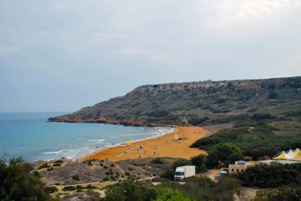 A sandy beach curves along the coastline with gentle waves, bordered by shrub-covered hills and cliffs. People and umbrellas dot the shore, while a few vehicles and tents near the entrance invite Gozo tourists to explore one of its hidden gems.