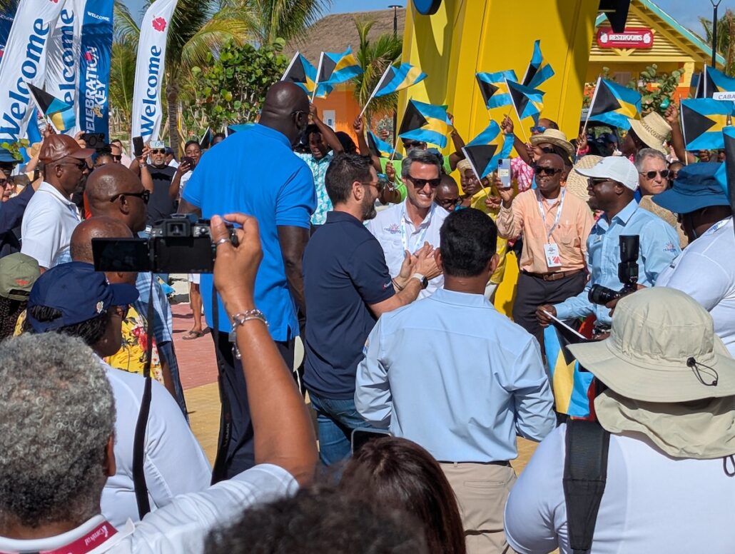 A crowd gathers outdoors near palm trees and a yellow sculpture for the Grand Opening, many holding small Bahamian flags. Some take photos while others wear lanyards, as "Welcome" banners mark the festive atmosphere at Celebration Key.