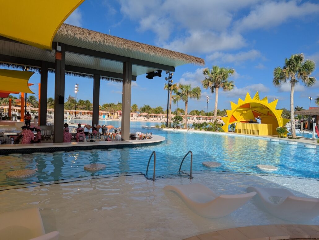 A large outdoor pool at Celebration Key features submerged lounge chairs, a swim-up bar, and a stage with a yellow sun decoration. Palm trees and people are visible under a blue sky with scattered clouds.