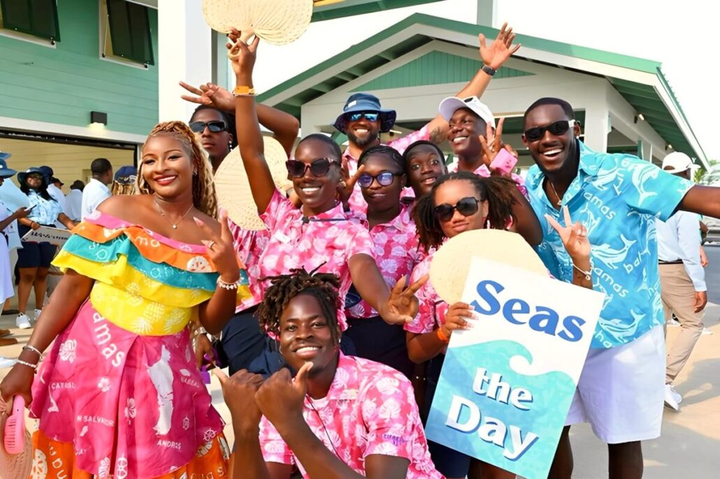 A group of people wearing colorful outfits and sunglasses pose and smile outdoors on a Locals Tour in Grand Bahama. One person holds a sign that reads “Seas the Day.” A green and white building is in the background.