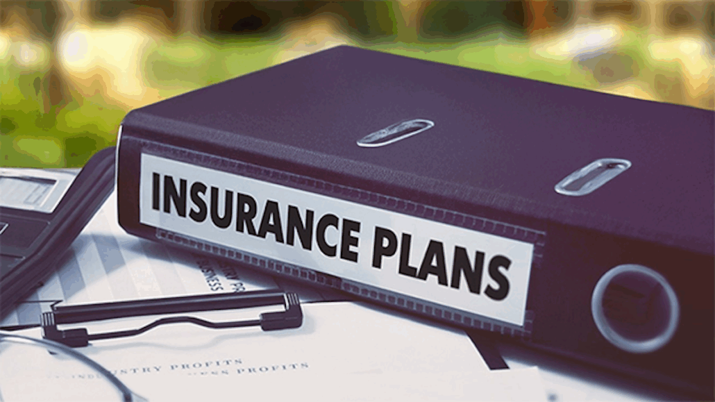 A close-up of a black binder labeled "Insurance Plans" resting on top of scattered papers and a clipboard, suggesting the importance of health coverage and travel insurance, with a blurred background.