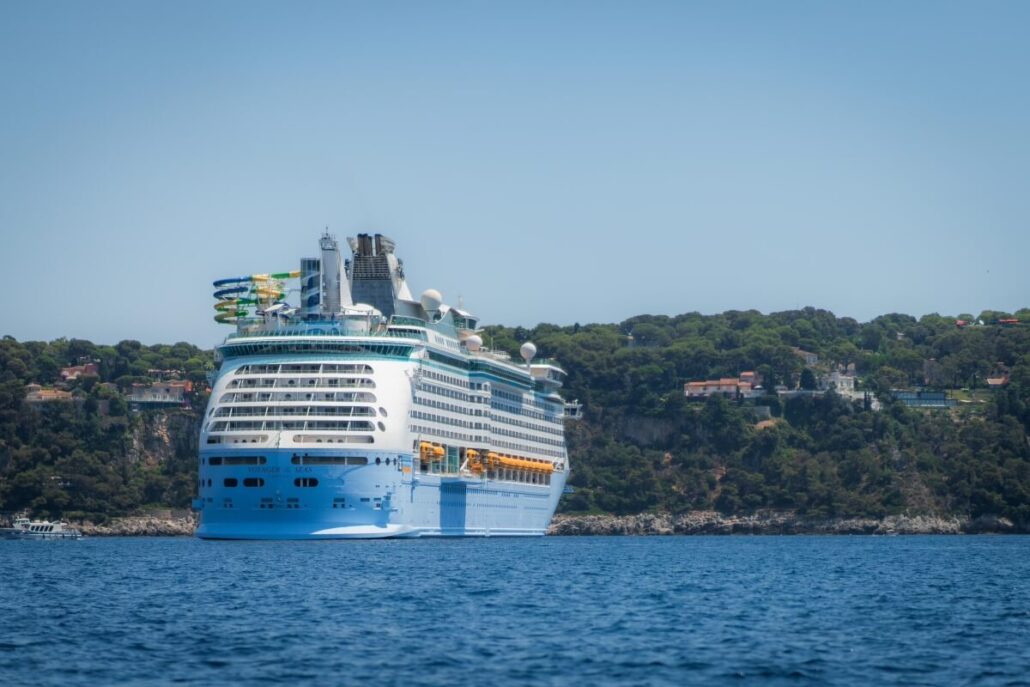 A large white and blue cruise ship is anchored near a forested coastline, with houses visible on the hillside in the background, under a clear blue sky—a scene reminiscent of the Mayor of Nice’s coastal jurisdiction.