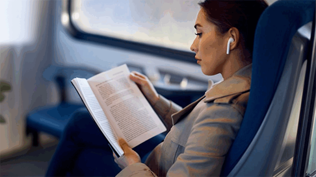 A woman wearing a beige coat sits in a train seat, reading a book and listening to wireless earbuds, feeling secure knowing she has travel insurance and health coverage protected abroad. The background shows a window with a blurred outdoor view.