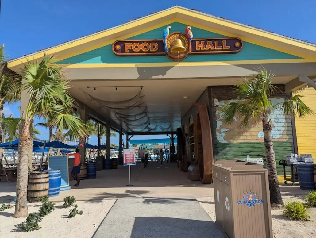 A building labeled "Food Hall" with a nautical theme stands surrounded by small palm trees and barrels. Tables with blue umbrellas and a Celebration Key trash bin set the scene for this vibrant Grand Opening.