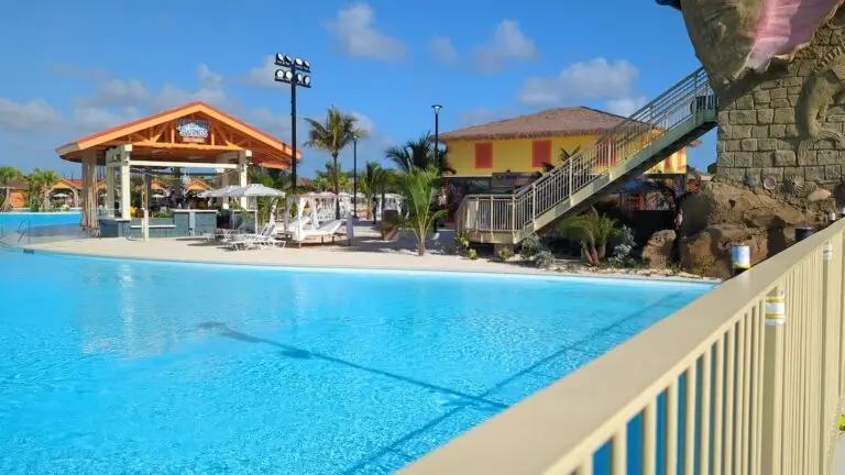 A swimming pool area with clear blue water, lounge chairs, umbrellas, a shaded bar, and a waterslide. There are palm trees and buildings in the background under a mostly sunny sky.
