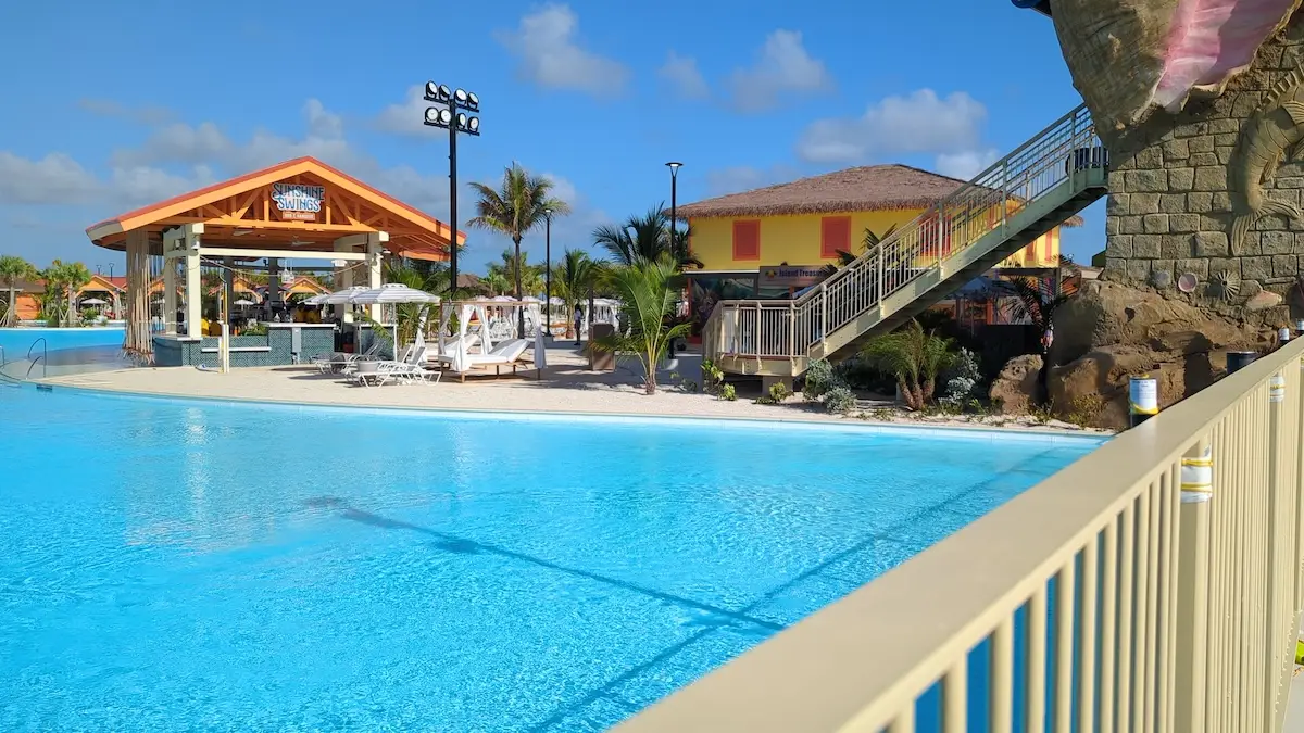 A swimming pool area with clear blue water, lounge chairs, umbrellas, a shaded bar, and a waterslide. There are palm trees and buildings in the background under a mostly sunny sky.