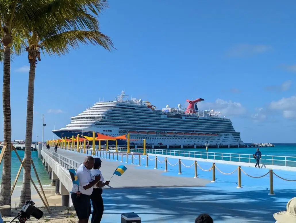 A large white cruise ship is docked at Celebration Key’s pier beside clear blue water under a sunny sky. Two people in the foreground hold small flags, and palm trees frame the left side of this grand opening scene.