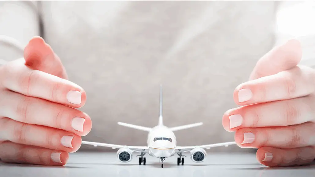 A close-up of a person’s hands cupping around a small model airplane on a white surface, giving the appearance of protecting or shielding the plane. The background and clothing are light in color.