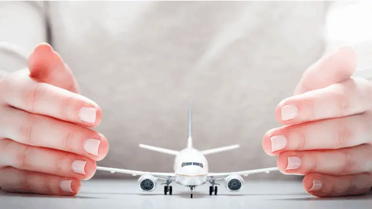 A close-up of a person’s hands cupping around a small model airplane on a white surface, giving the appearance of protecting or shielding the plane. The background and clothing are light in color.
