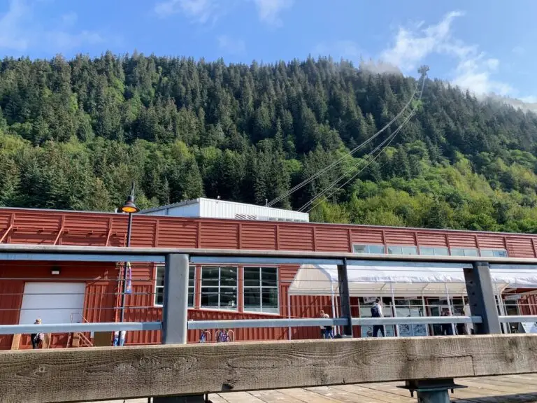 A red building with large windows stands before a forested hillside, where cables lead up to a tram station. In the foreground, a wooden railing and pier evoke the sense of adventure often found on an Alaska cruise.