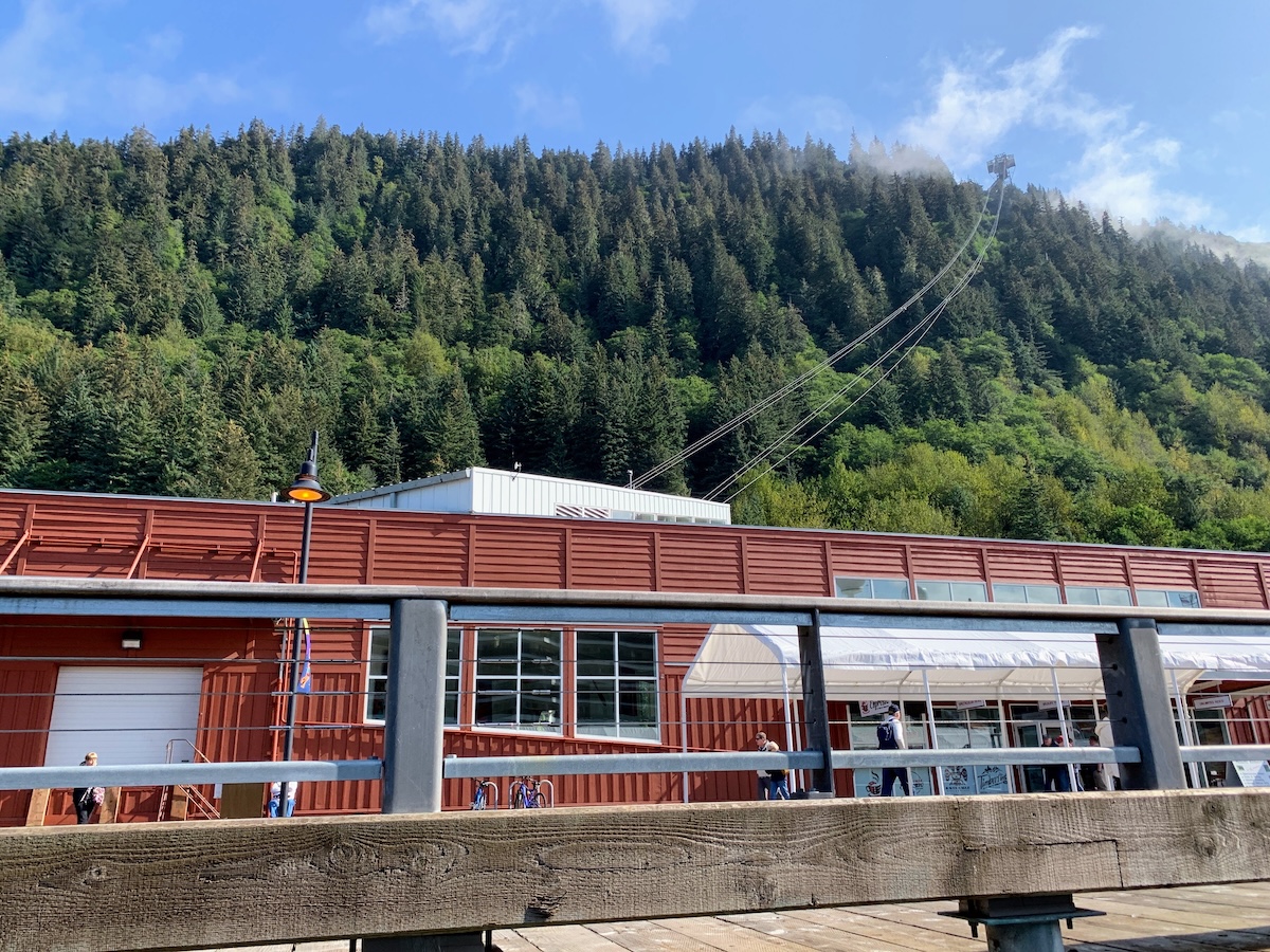 A red building with large windows stands before a forested hillside, where cables lead up to a tram station. In the foreground, a wooden railing and pier evoke the sense of adventure often found on an Alaska cruise.
