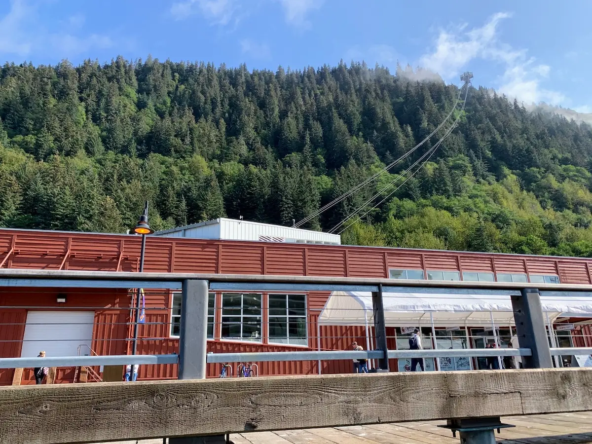A red building with large windows stands before a forested hillside, where cables lead up to a tram station. In the foreground, a wooden railing and pier evoke the sense of adventure often found on an Alaska cruise.