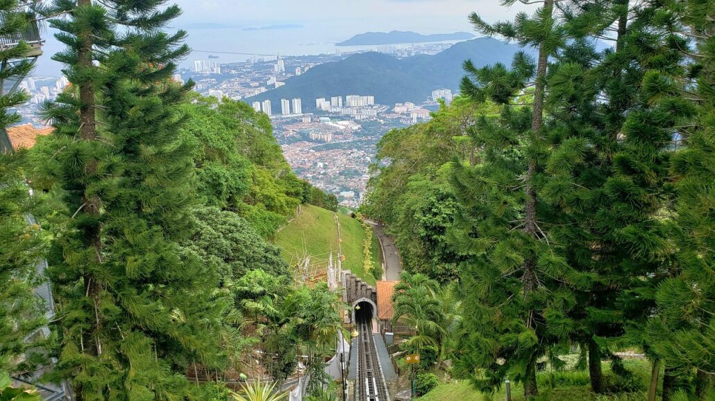 A funicular railway track runs down a green hillside lined with tall trees, leading to a tunnel entrance. In the background, Attractions like TeddyVille and Magic World enhance the cityscape and coastline beneath a partly cloudy sky.