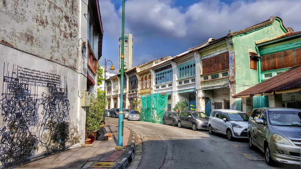 A street lined with old, colorful shophouses—some restored as unique attractions—features parked cars, a streetlamp, and wall art on a building beneath a partly cloudy blue sky.