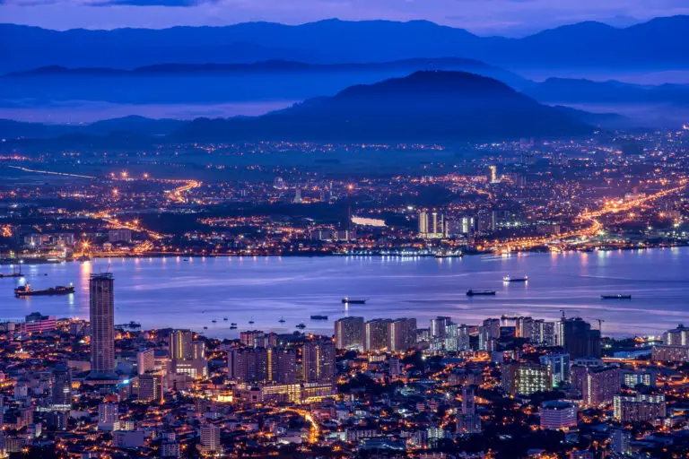A cityscape at dusk shows illuminated buildings near a waterfront, with boats in the water and mountains in the background under a blue and purple sky. Magic World attractions and streetlights add vibrance to the scene, with lights glowing throughout.