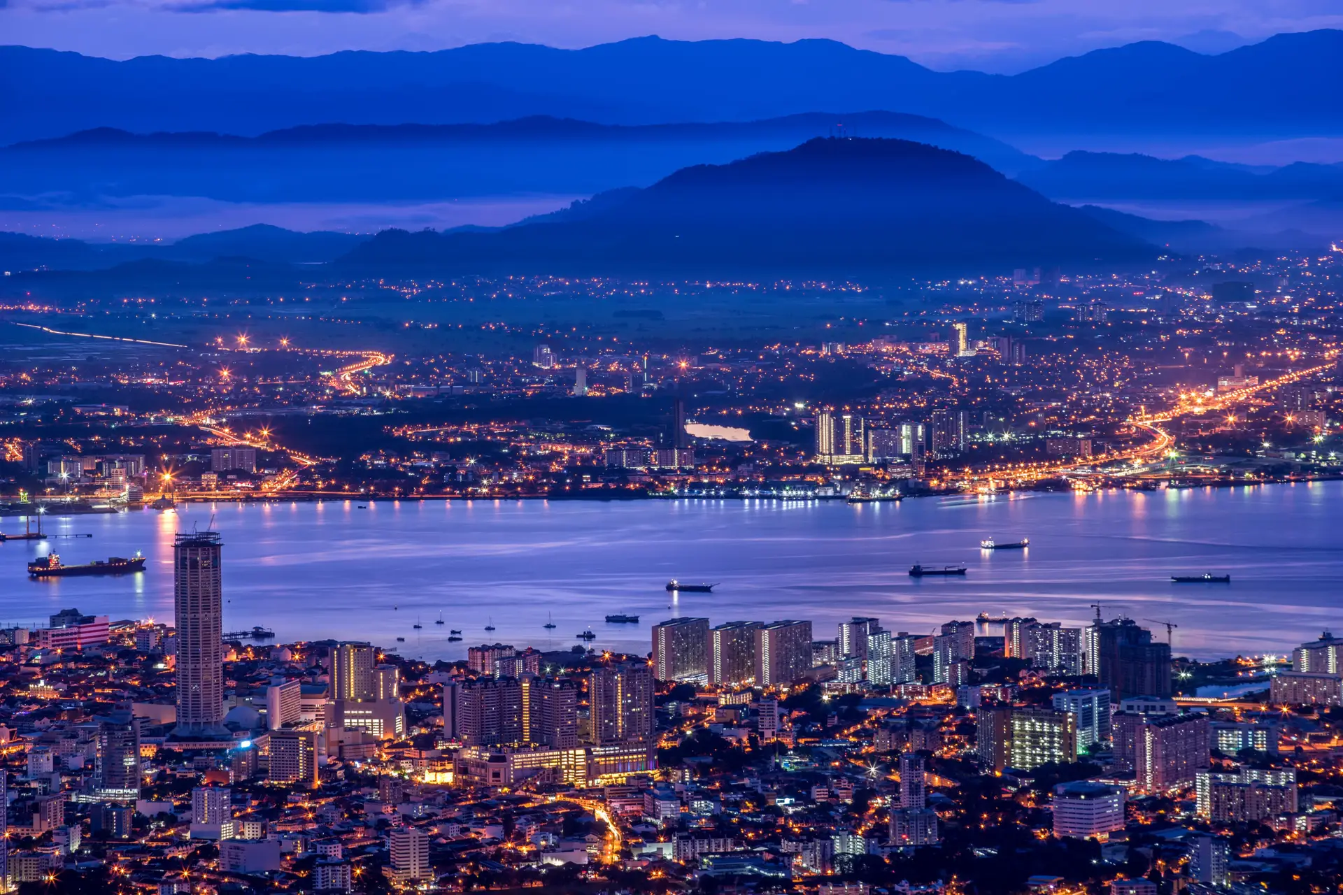 A cityscape at dusk shows illuminated buildings near a waterfront, with boats in the water and mountains in the background under a blue and purple sky. Magic World attractions and streetlights add vibrance to the scene, with lights glowing throughout.