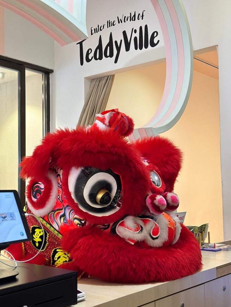 A large red lion dance costume with white, black, and gold details is displayed on a counter beneath a sign that reads "Enter the World of TeddyVille," inviting visitors to explore one of TeddyVille’s enchanting attractions. The costume has bold eyes and pink accents.