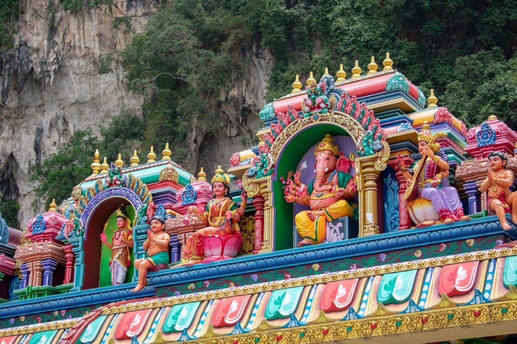 Colorful statues of Hindu deities and figures adorn the ornate, detailed rooftop of this temple, making it one of the must-see attractions, with a rocky cliff and lush greenery providing a stunning backdrop.