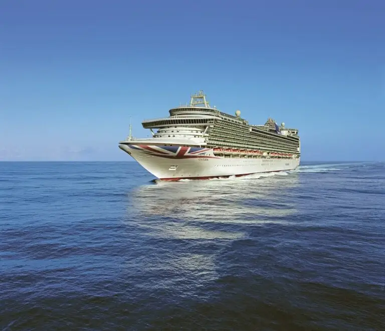 A large white cruise ship with a painted Union Jack on the bow sails on calm blue ocean water under a clear sky.