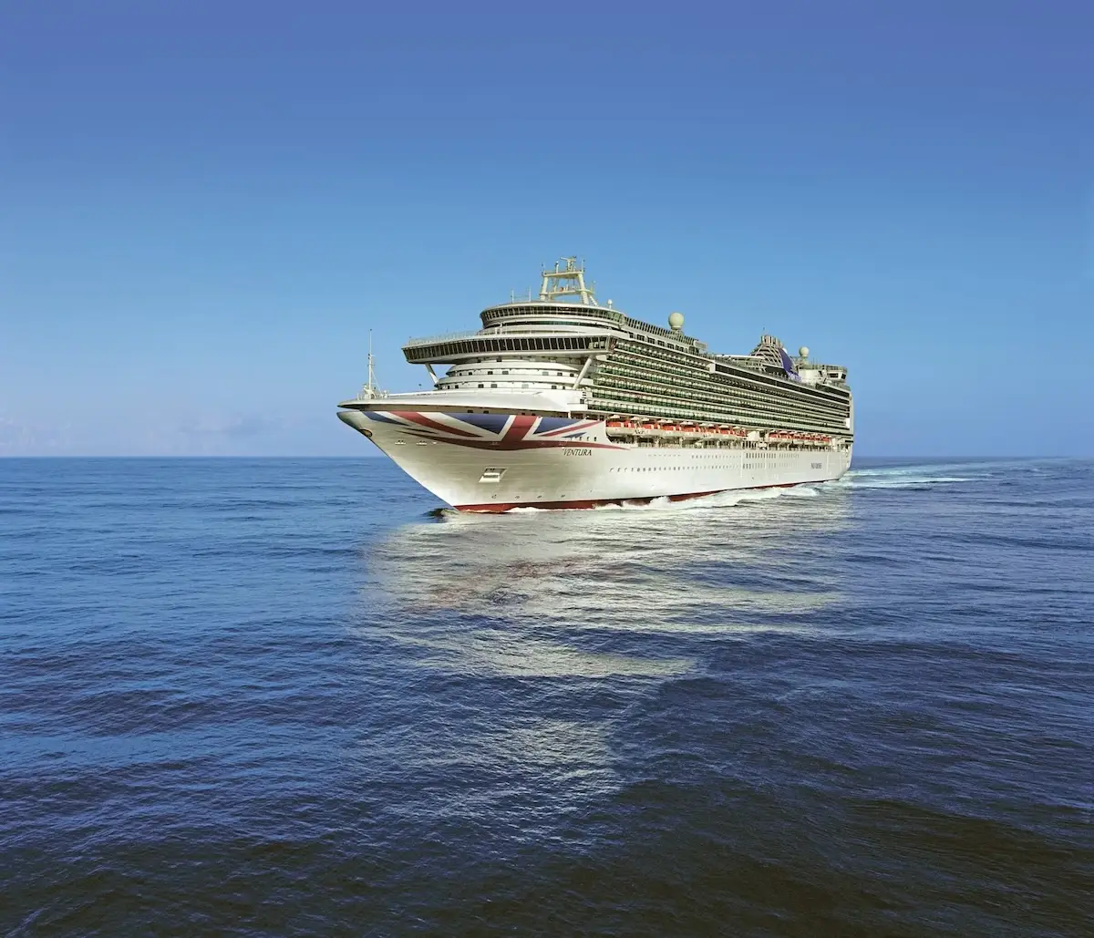 A large white cruise ship with a painted Union Jack on the bow sails on calm blue ocean water under a clear sky.