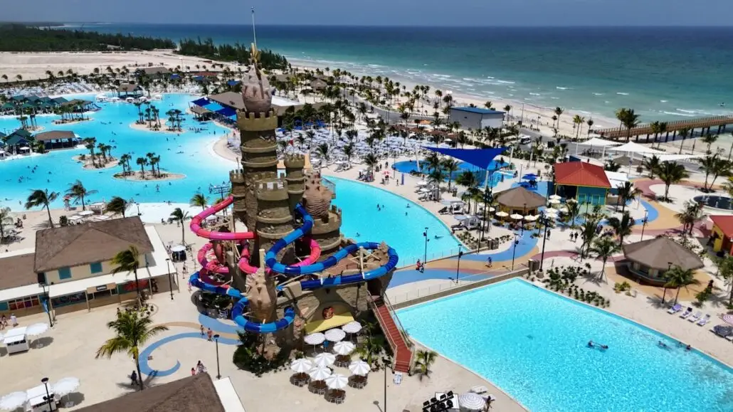 A large water park by the ocean, near the Carnival Celebration Key, features several pools, palm trees, lounge chairs, and a central waterslide tower shaped like a sandcastle with colorful slides. The beach and sea are visible in the background.
