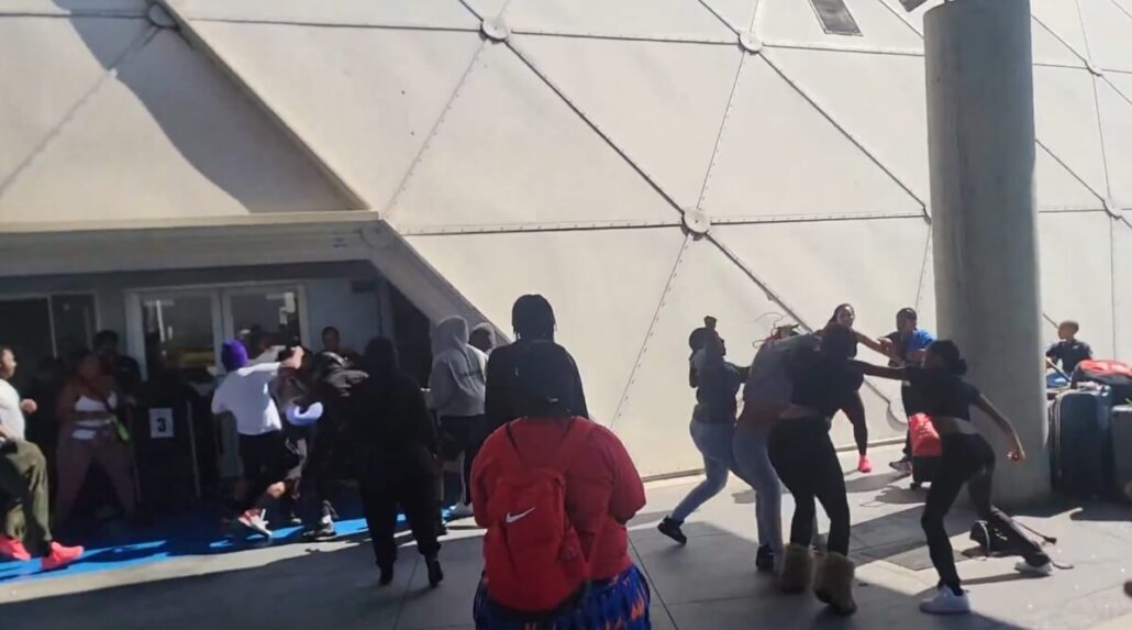 A group of people, including some Carnival passengers, are involved in a physical altercation outside a building with a geometric wall near the Long Beach Cruise Terminal. Some are fighting, while others observe or move away. A large concrete pillar is on the right.