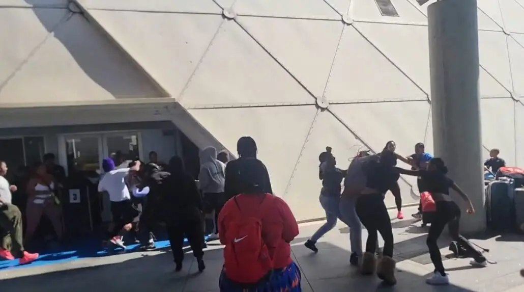 A group of people, including some Carnival passengers, are involved in a physical altercation outside a building with a geometric wall near the Long Beach Cruise Terminal. Some are fighting, while others observe or move away. A large concrete pillar is on the right.