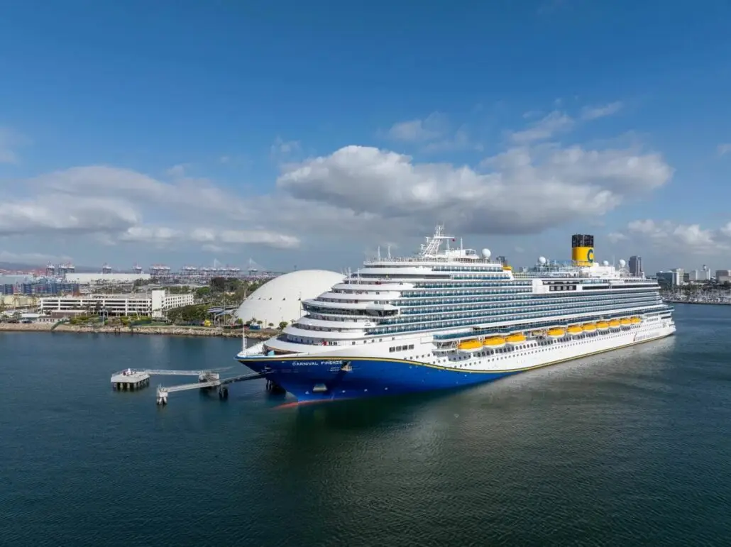 A large cruise ship with a blue hull and yellow lifeboats is docked at the Long Beach Cruise Terminal beside a white domed building, with city buildings and cranes visible in the background under a partly cloudy sky.