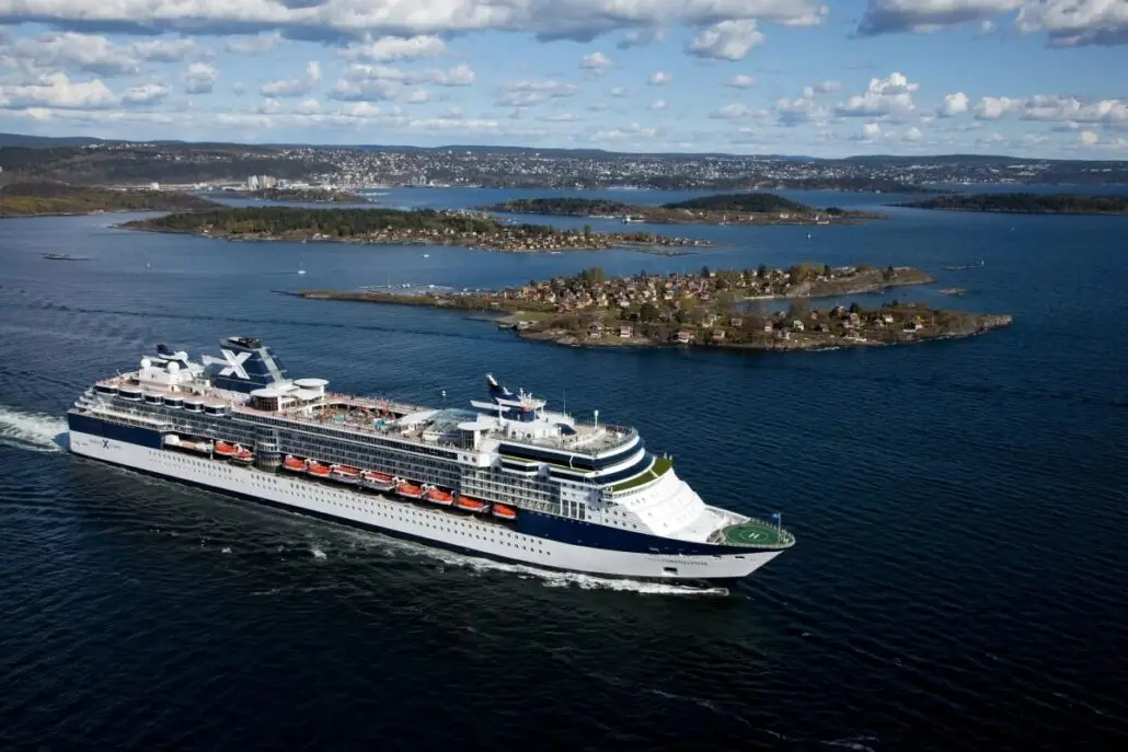 A large Celebrity Cruises ship sails on a calm body of water near several small, green islands with houses, under a partly cloudy sky. The distant Italy coast with shoreline buildings is visible in the background.