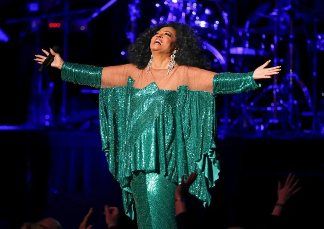 A performer in a sparkly green dress stands on stage with arms outstretched, smiling and holding a microphone—channeling Diana Ross vibes aboard the exciting new Royal Caribbean ship. Stage lights and musical instruments shine in the background.