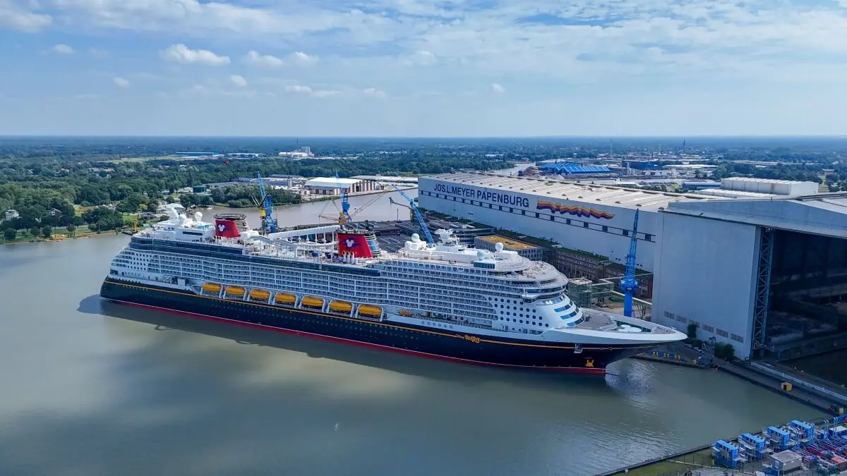 A large cruise ship, Disney Destiny, is docked beside the Meyer Werft Papenburg shipyard on a calm river, with trees and industrial buildings in the background under a partly cloudy sky ahead of its November Launch.