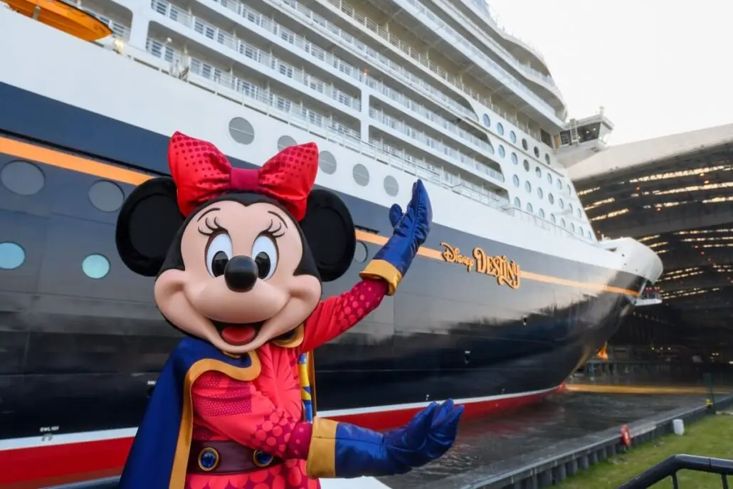 A person in a Minnie Mouse costume stands in front of the Disney Destiny cruise ship, built by Meyer Werft and set for its November Launch. Minnie Mouse poses with both arms raised, gesturing proudly toward the impressive ship.