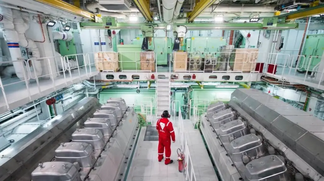 A cruise ship engineer in red coveralls and a helmet stands in the middle of an industrial engine room with large machinery, pipes, and control panels on both sides and multiple levels visible.