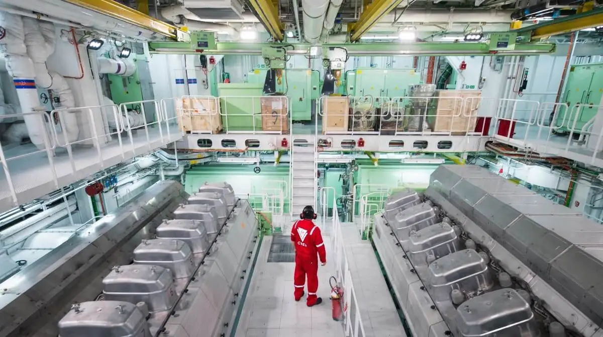 A cruise ship engineer in red coveralls and a helmet stands in the middle of an industrial engine room with large machinery, pipes, and control panels on both sides and multiple levels visible.