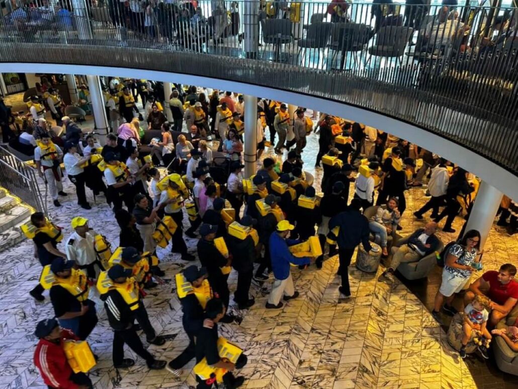 A large group of people, many wearing yellow life jackets and hats, gather indoors on a marble floor, as cruise ship news circulates. Some are seated while others stand or walk, with more people visible above on the balcony.